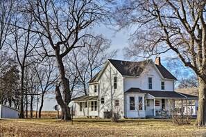 Una casa bianca a due piani con un portico, circondata da alberi e un cielo sereno.