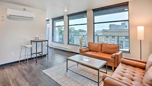 A modern living room with a leather sofa, a coffee table, and a dining area with a chair and table. Large windows provide a view of the cityscape. - Lake Norden Apartment w/ Spacious Balcony! (Lake Norden)