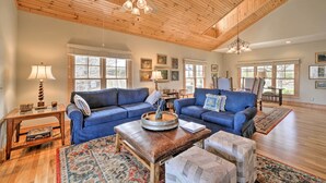 A spacious living room with high ceilings, wooden beams, and large windows. There are two blue sofas, a coffee table, and a rug. - Beautiful Lake Glenville Townhome w/ Mountain View (Cashiers)