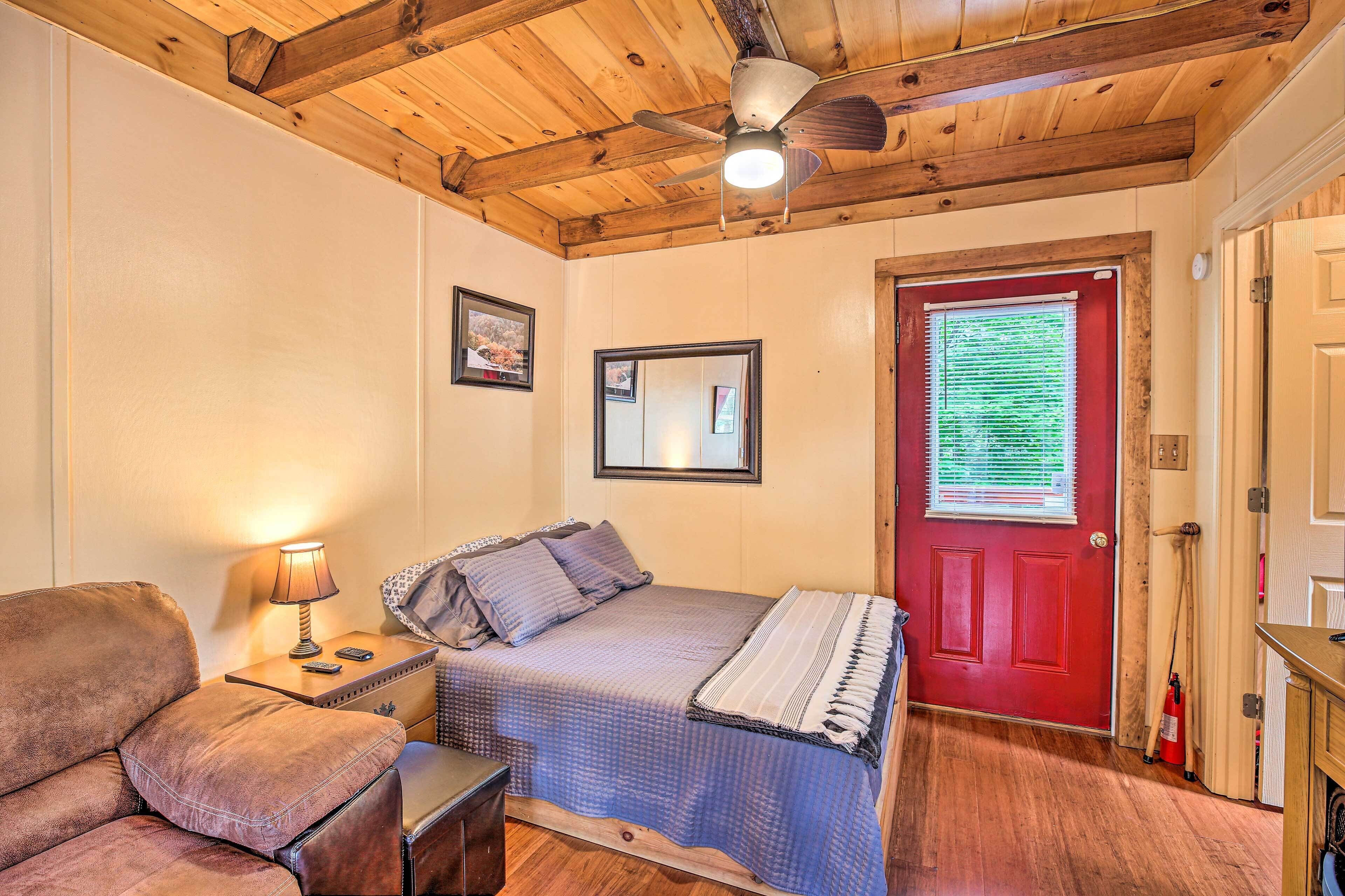 A bedroom with a bed, a nightstand, a lamp, a wooden ceiling, a red door, and a window with blinds.