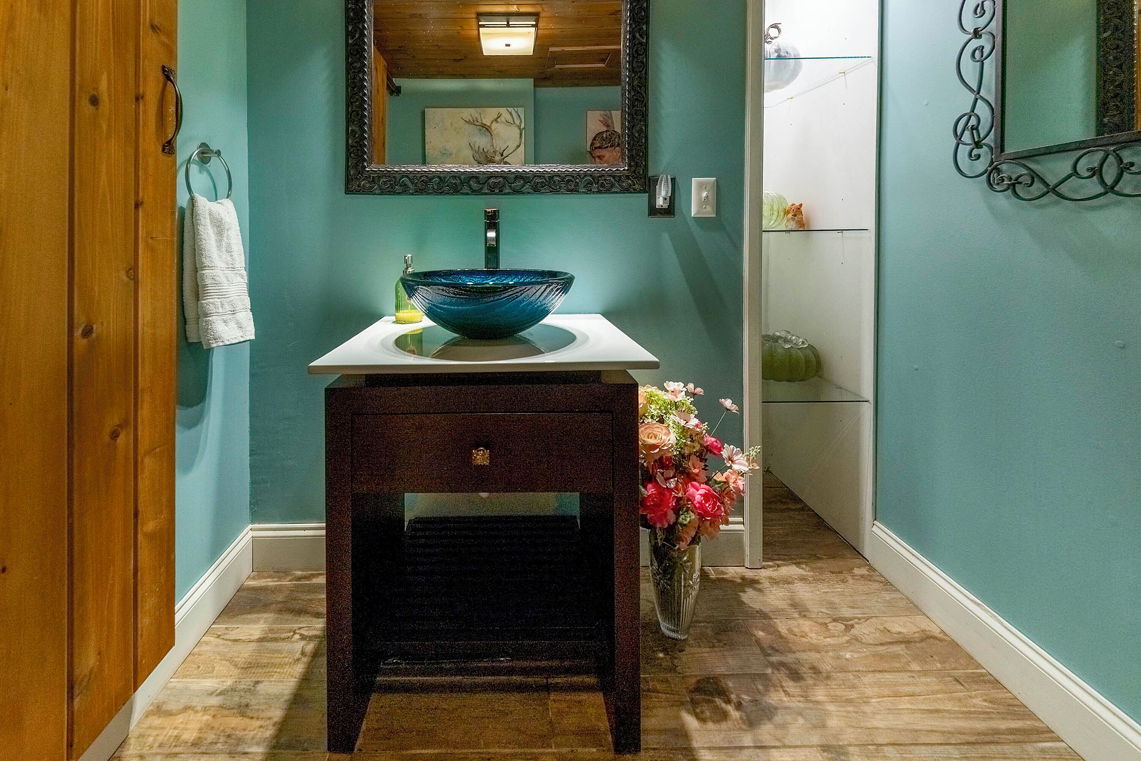Une salle de bain avec un lavabo en verre bleu, un évier blanc, un meuble-lavabo en bois et un vase rempli de fleurs.