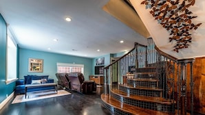 A spacious living area with a dark wooden floor, a blue sofa, a leather armchair, and a staircase with a decorative railing. - Woodland Spa Sanctuary: Delaware Home w/ Swim Spa (Delaware)