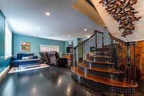 Uma sala de estar ampla com piso de madeira escura, um sofá azul, uma poltrona de couro e uma escada com corrimão decorativo.