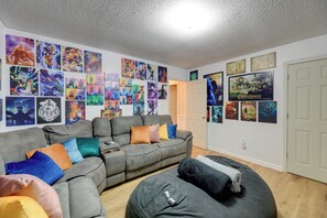 A living room with a grey sofa, colorful cushions, and a bean bag chair. The walls are decorated with anime and movie posters.