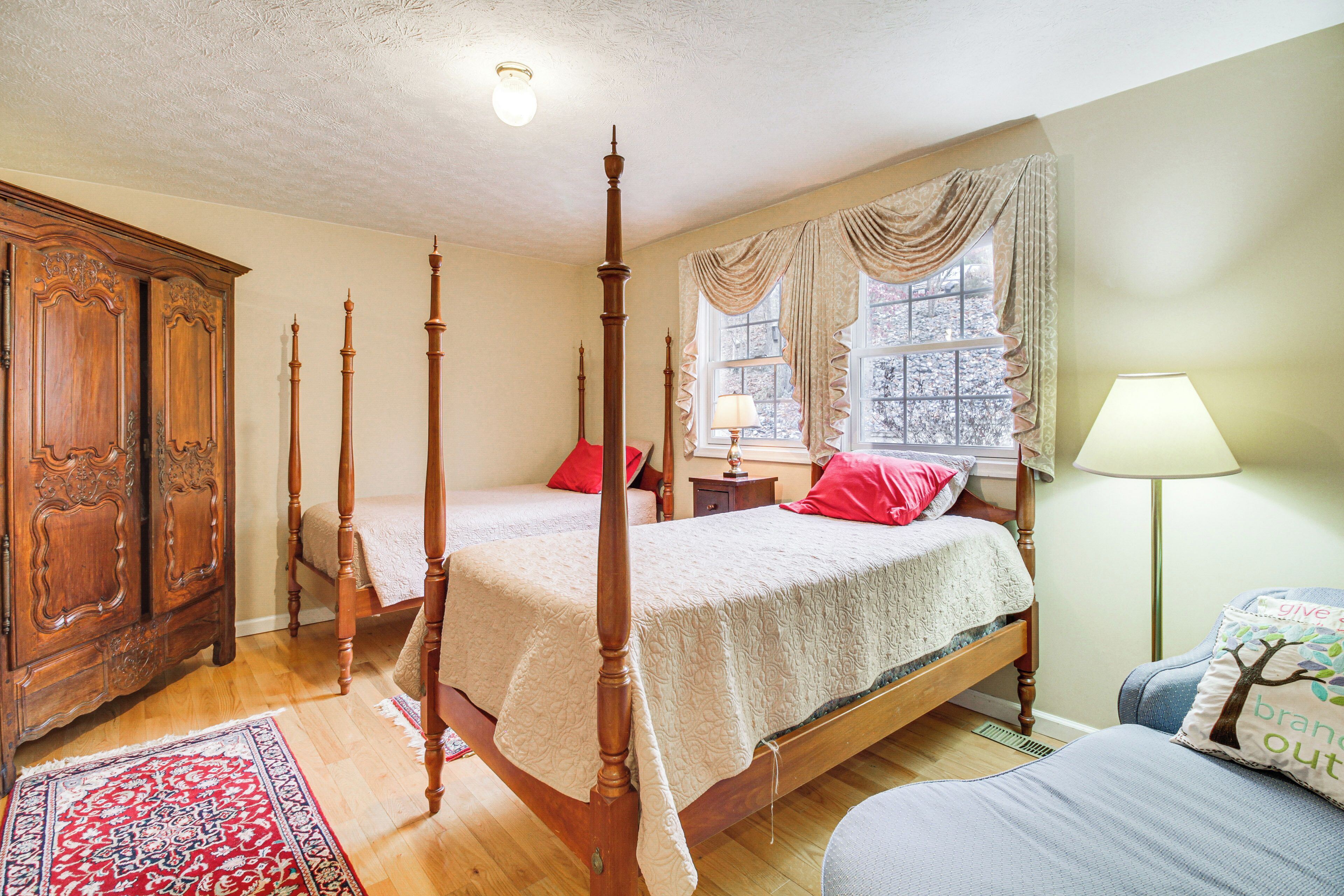 Un lit à baldaquin avec un cadre en bois, deux tables de chevet et une petite table d’appoint. Il y a une fenêtre avec des rideaux, une lampe de sol et un tapis décoratif.
