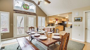 A dining area with a large wooden table set for six, a ceiling fan, and a view of the kitchen and living areas through large windows. - Resort Home on Mountain Golf Course w/ Deck & View (McGaheysville)