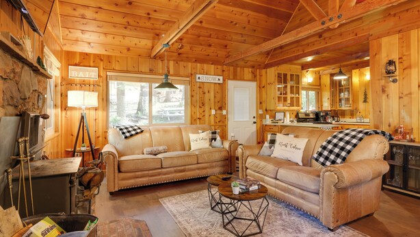 A cozy cabin interior with wooden walls and ceilings, two leather sofas with checkered throws, a dining table, and a stone fireplace. - 3 Mi to Big Trees State Park: Cabin w/ Scenic Deck (Arnold)