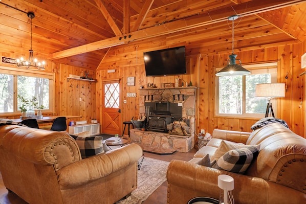 A cozy log cabin interior with leather sofas, a stone fireplace, a television, and wooden walls. - 3 Mi to Big Trees State Park: Cabin w/ Scenic Deck (Arnold)