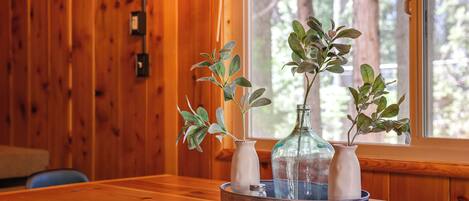 A wooden interior with a round metal tray holding three white vases and a glass bottle, a window with a view of trees, and a wooden wall with a light switch.