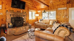 A cozy cabin interior with a stone fireplace, leather sofa, and a television mounted on the wall. - 3 Mi to Big Trees State Park: Cabin w/ Scenic Deck (Arnold)