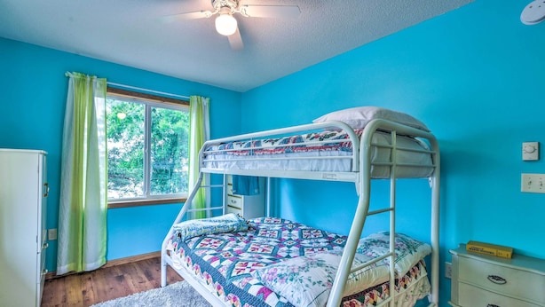 A bunk bed in a room with blue walls, a window with green curtains, a ceiling fan, and a small white dresser. - Lakefront Spicer Cabin w/ Dock & 2 Kayaks! (Spicer)
