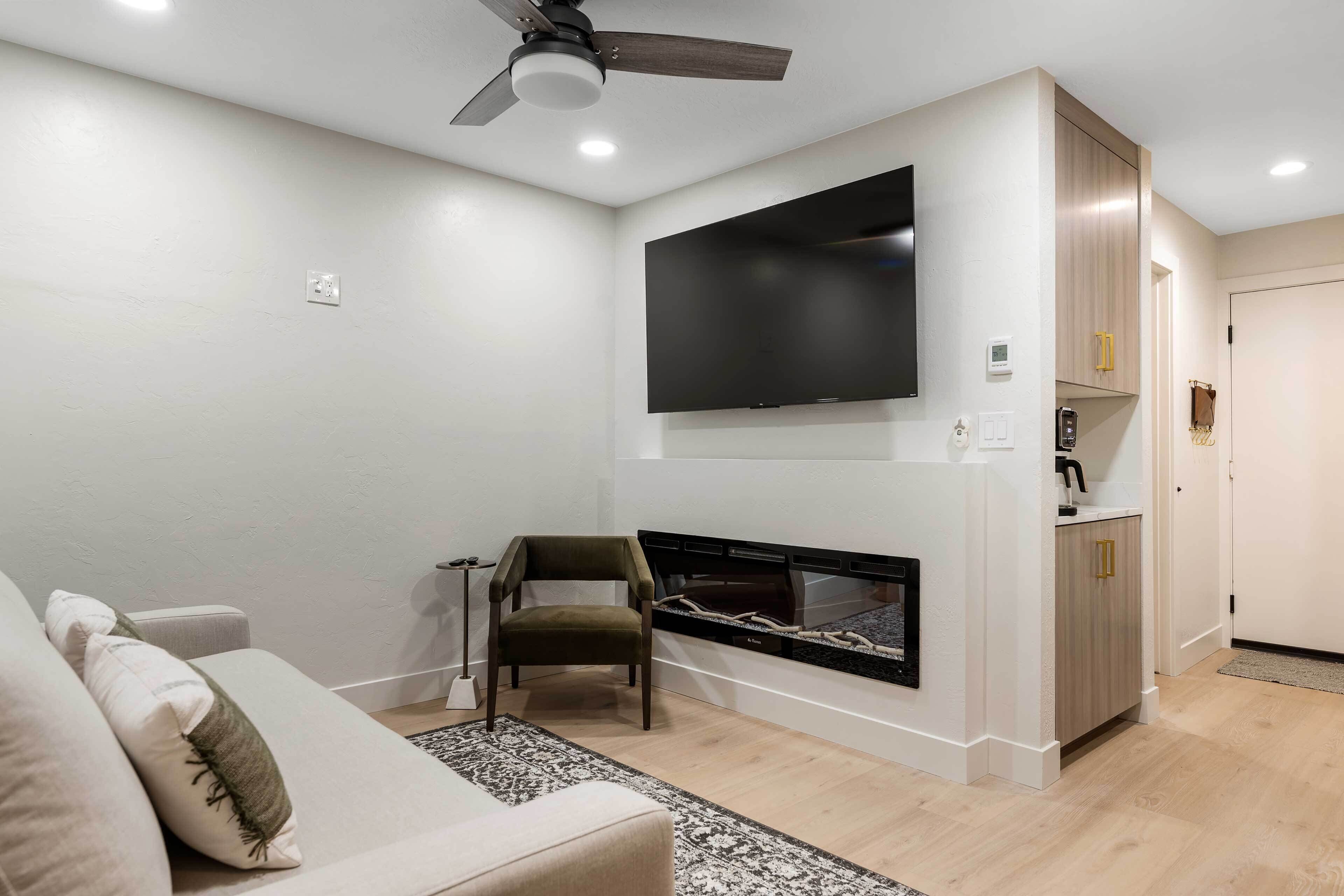 A living room with a sofa, a chair, a fireplace, a television mounted on the wall, and a ceiling fan.