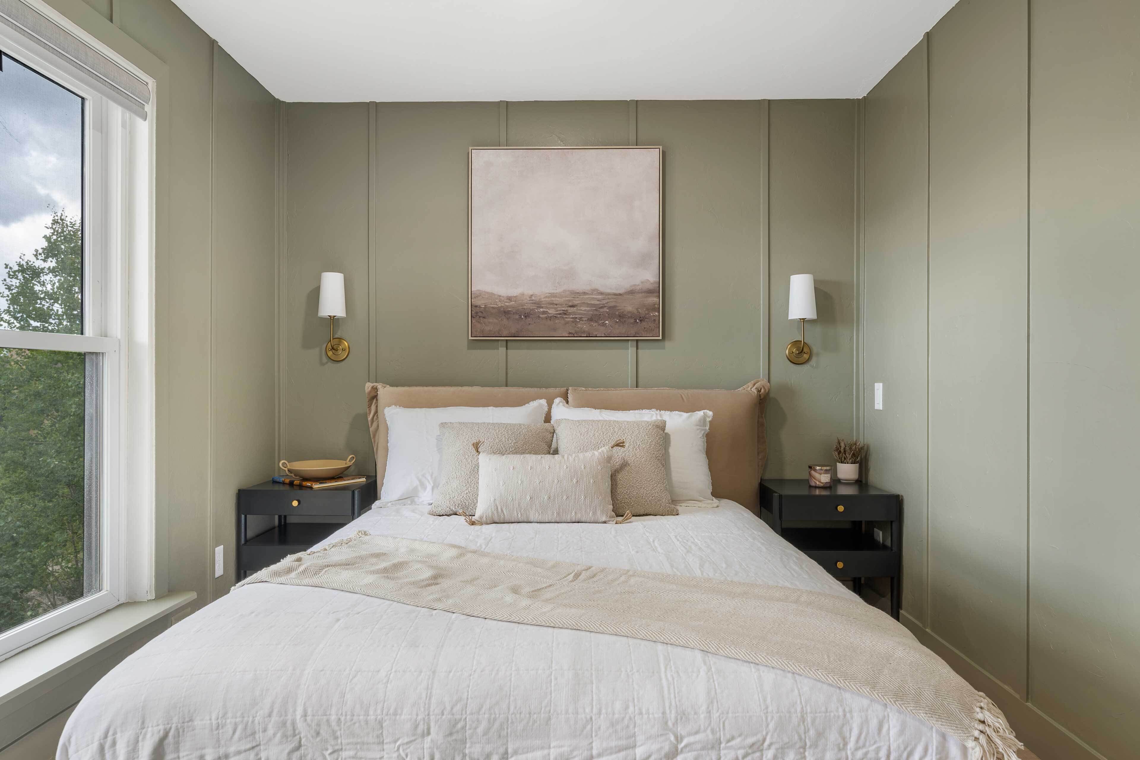 A bedroom with a large bed, two bedside tables, a window, and a wall-mounted painting.