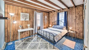 A bedroom with a wooden paneled wall, a bed with a metal frame, a bedside table with a lamp, a wall-mounted dreamcatcher, and a window with curtains. - Pet-friendly Mohnton House Near Golf Course! (Mohnton)