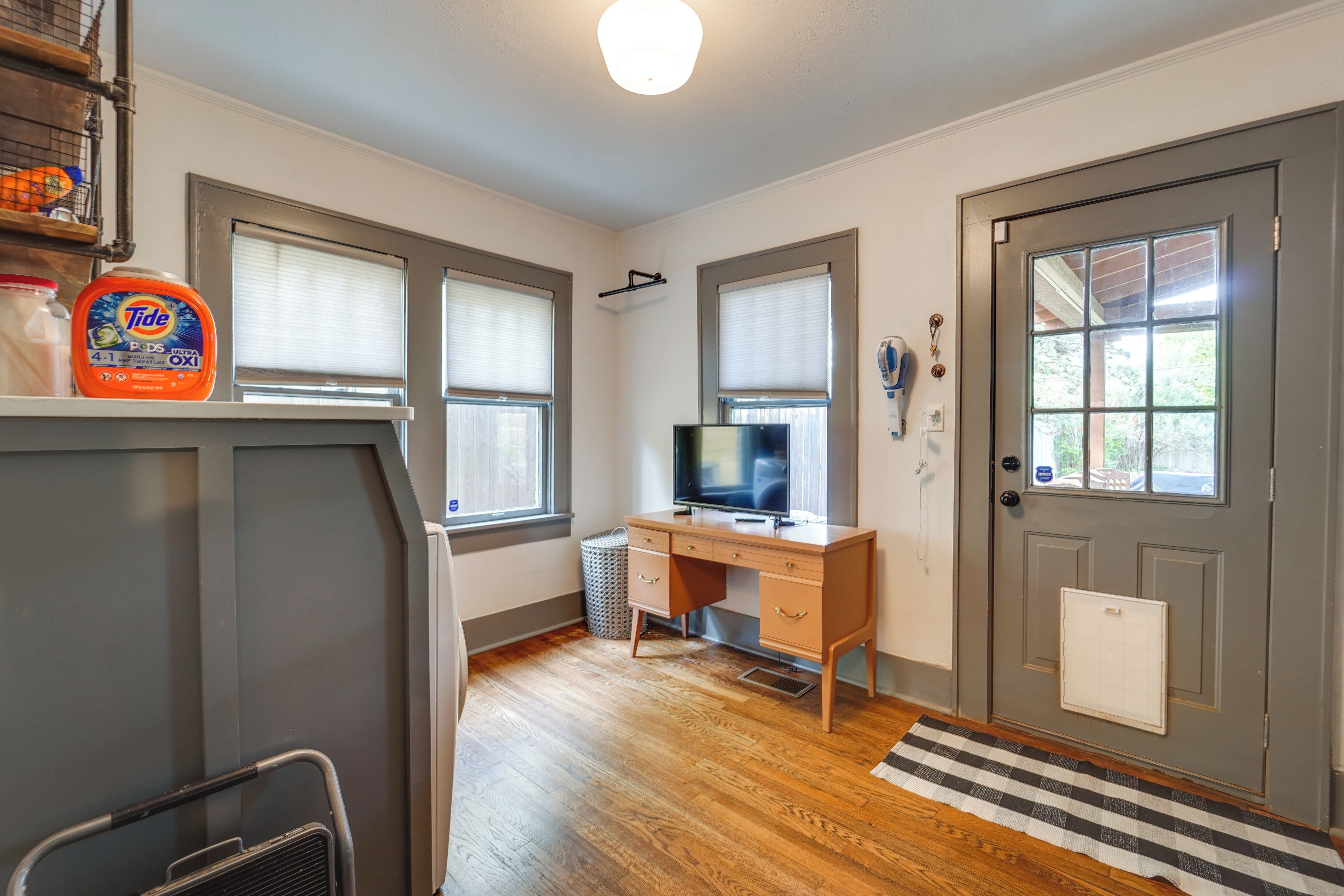 A room with a wooden desk, a television, a window with blinds, a door with a calendar, and a rug.