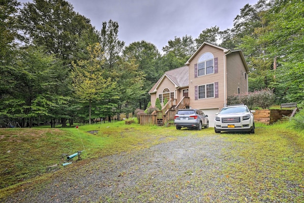 A two-story house with a driveway leading to a garage, surrounded by lush greenery and trees.
