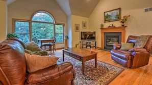 A spacious living room with a fireplace, leather sofas, a wooden coffee table, and a large window offering a view of the outdoors. - Pocono Mountain Home w/ 3 Community Lakes! (Gouldsboro)
