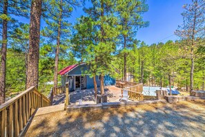 A cabin with a red roof, surrounded by tall pine trees, and a wooden deck with chairs. - Relaxing Ruidoso Getaway w/ Spacious Deck! (Ruidoso)