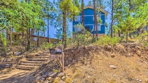 A building with a blue exterior, surrounded by trees and a dirt path leading to a wooden staircase. - Relaxing Ruidoso Getaway w/ Spacious Deck! (Ruidoso)