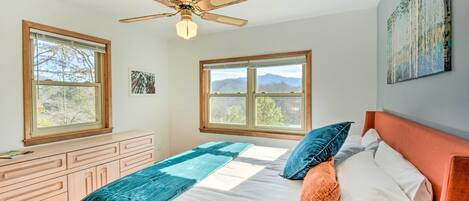 Ein Schlafzimmer mit einem Bett, einem Deckenventilator, zwei Fenstern, einem Schminktisch und einem Bild an der Wand.