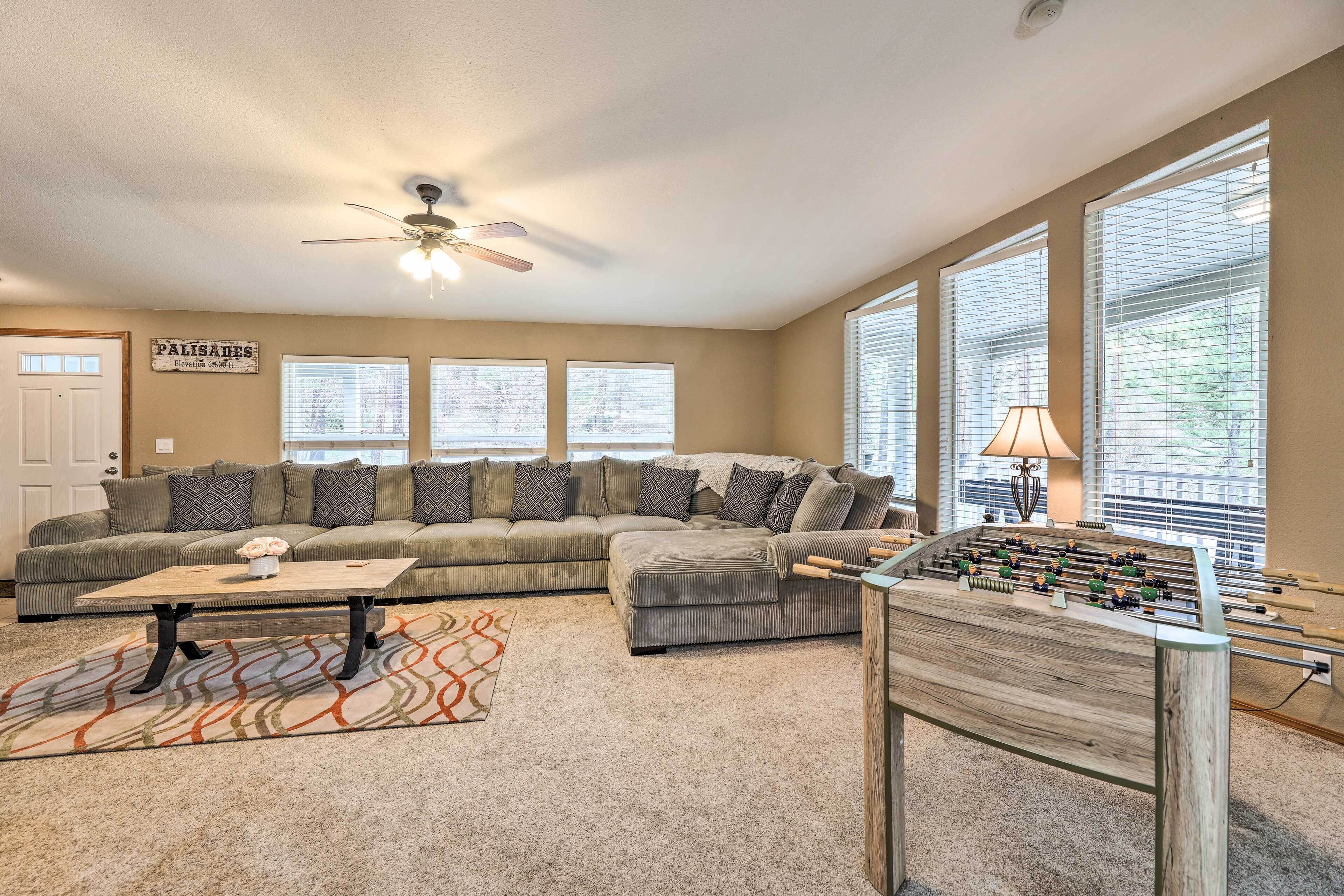 A living room with a sectional sofa, a coffee table, a foosball table, and large windows with blinds.