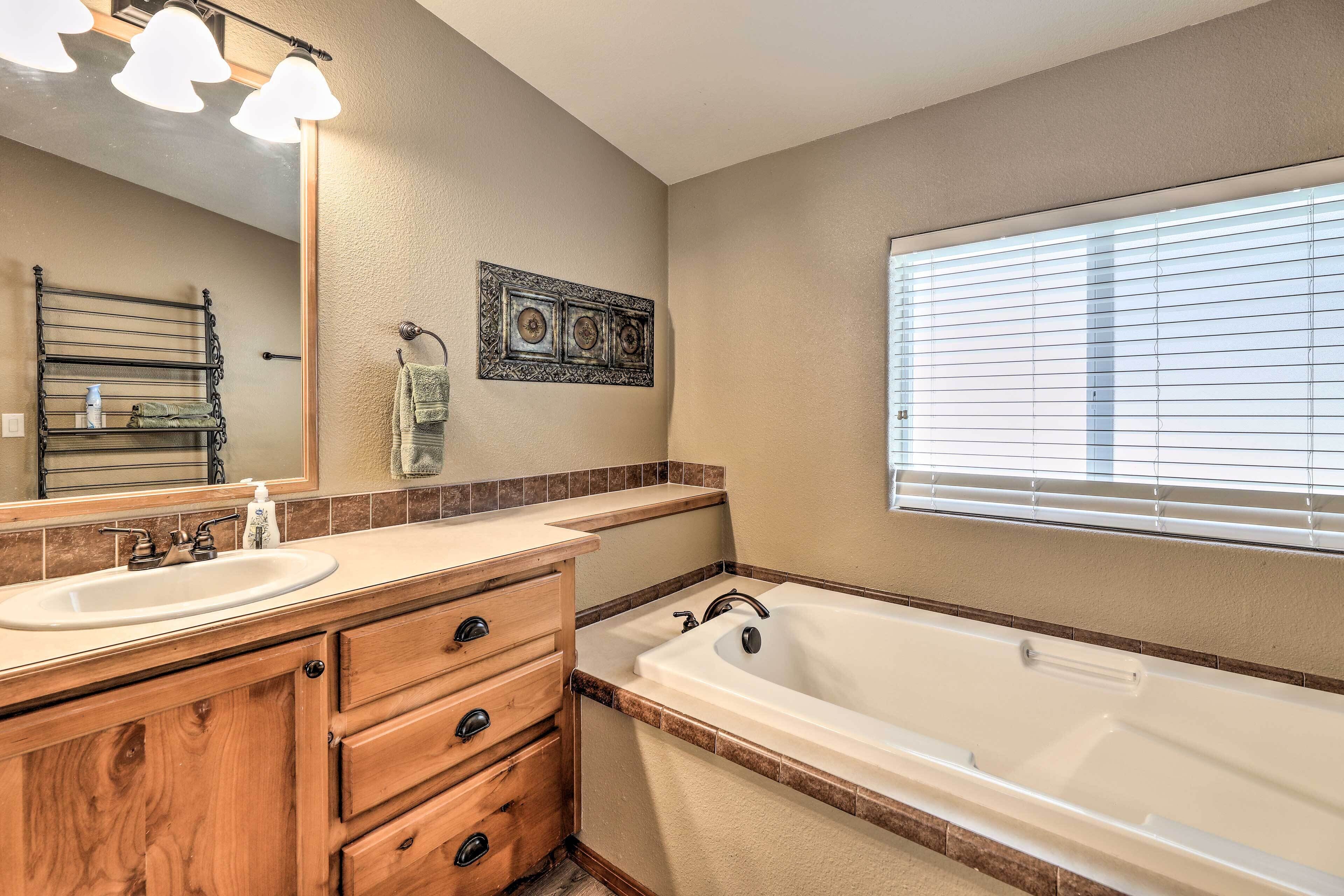 A bathroom with a wooden vanity, a large bathtub, a mirror, and a window with blinds.