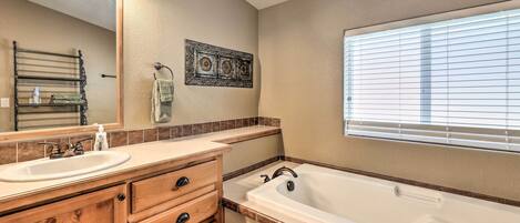 A bathroom with a wooden vanity, a large bathtub, a mirror, and a window with blinds.