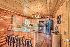 A wooden cabin kitchen with a central island, bar stools, stainless steel appliances, and a rustic ceiling.