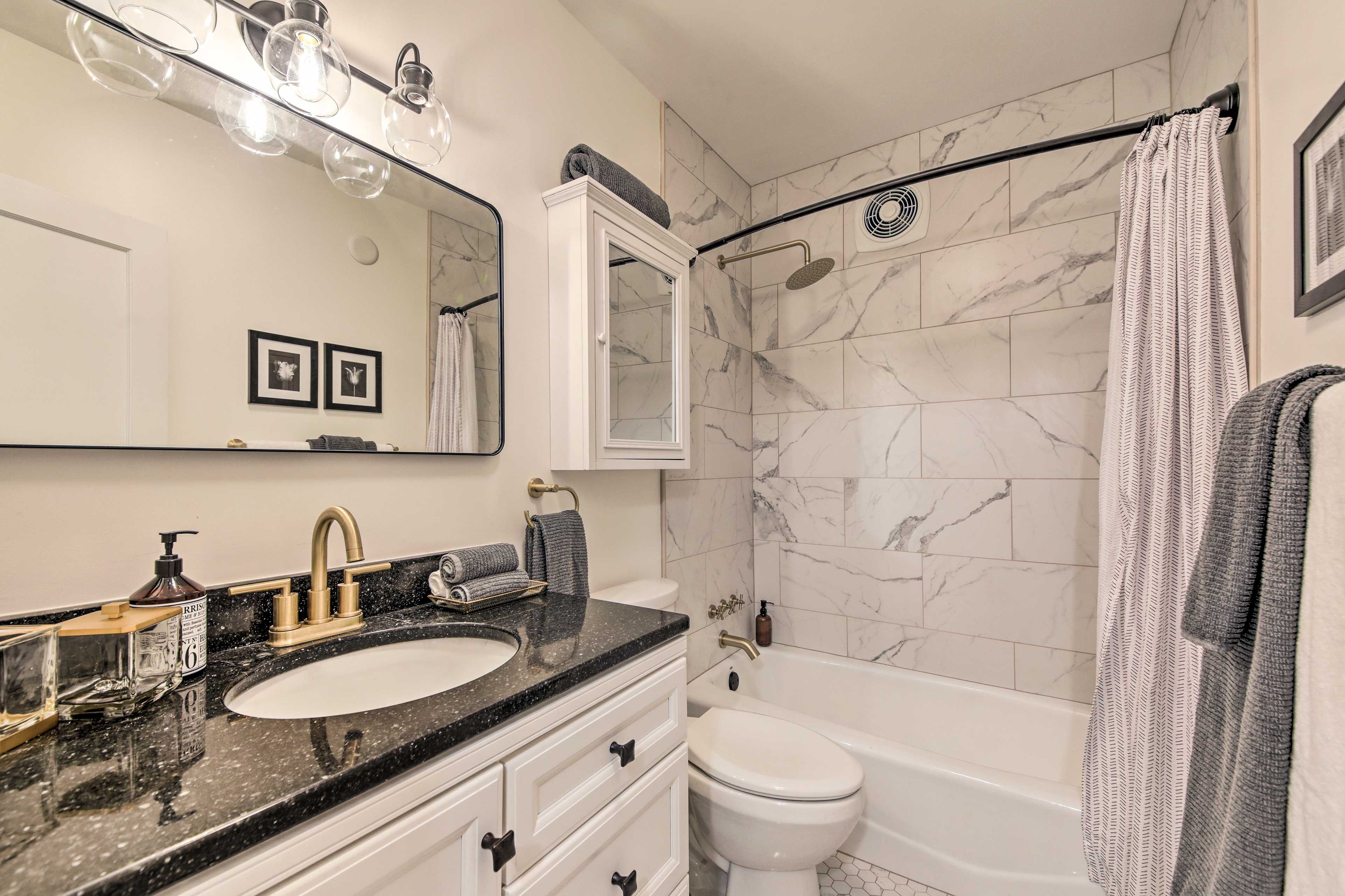 A bathroom with a marble-tiled shower, a white sink with a black countertop, and a large mirror with lights.