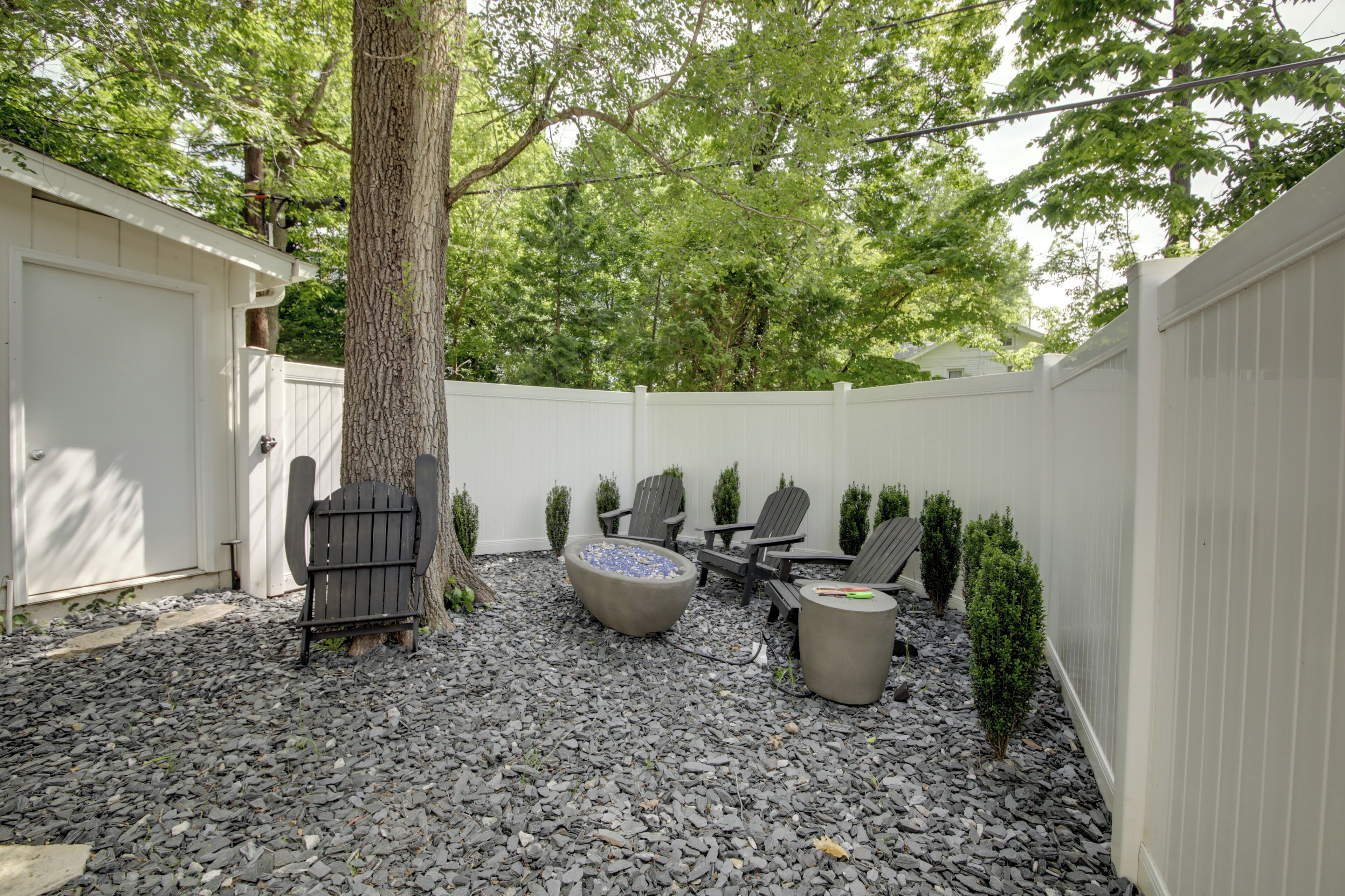 Un patio trasero con cerca blanca, un árbol, sillas Adirondack, un pozo de fuego y una pequeña zona de jardín.