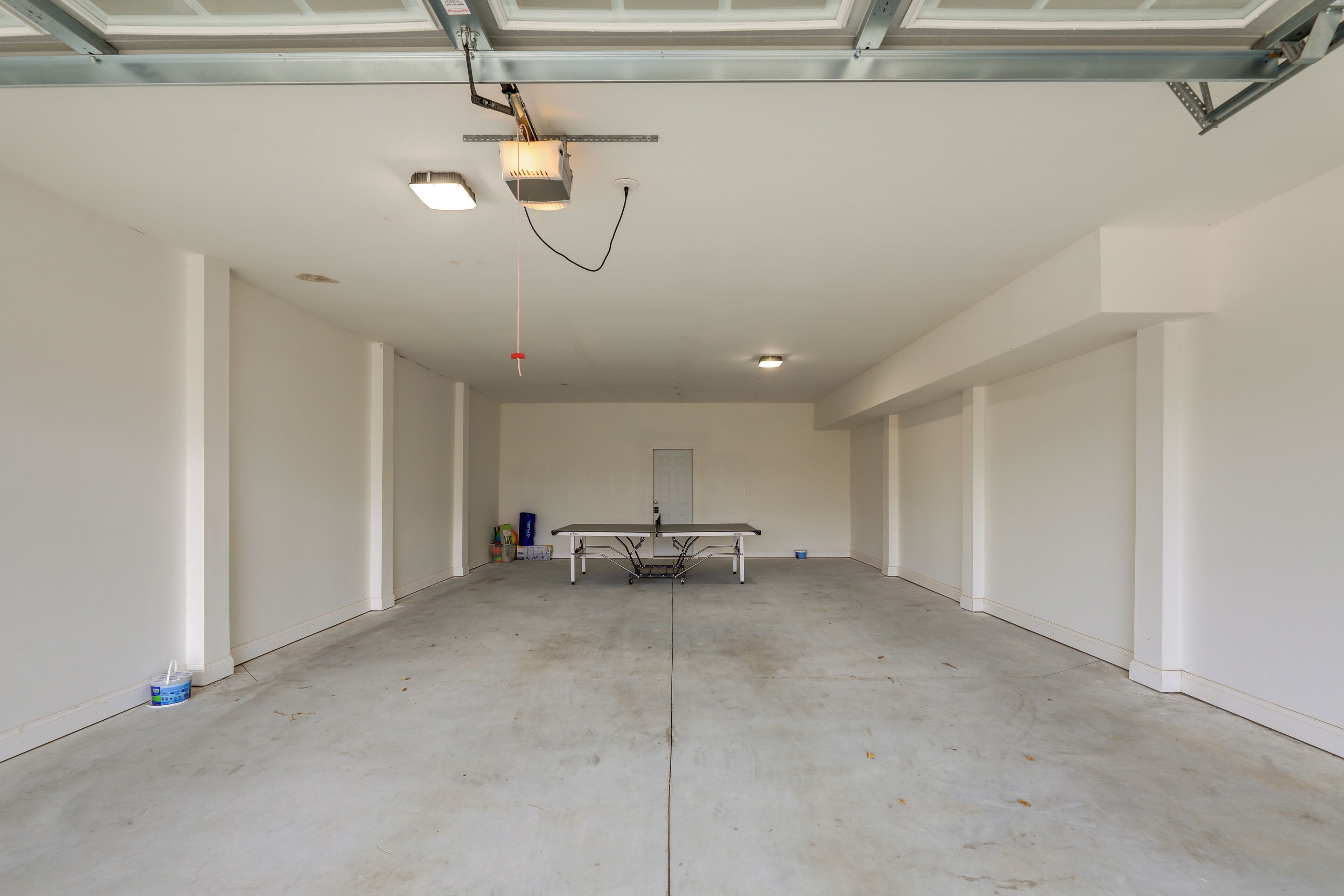 A spacious, empty room with a large table in the center, a ceiling-mounted projector, and a door at the far end.