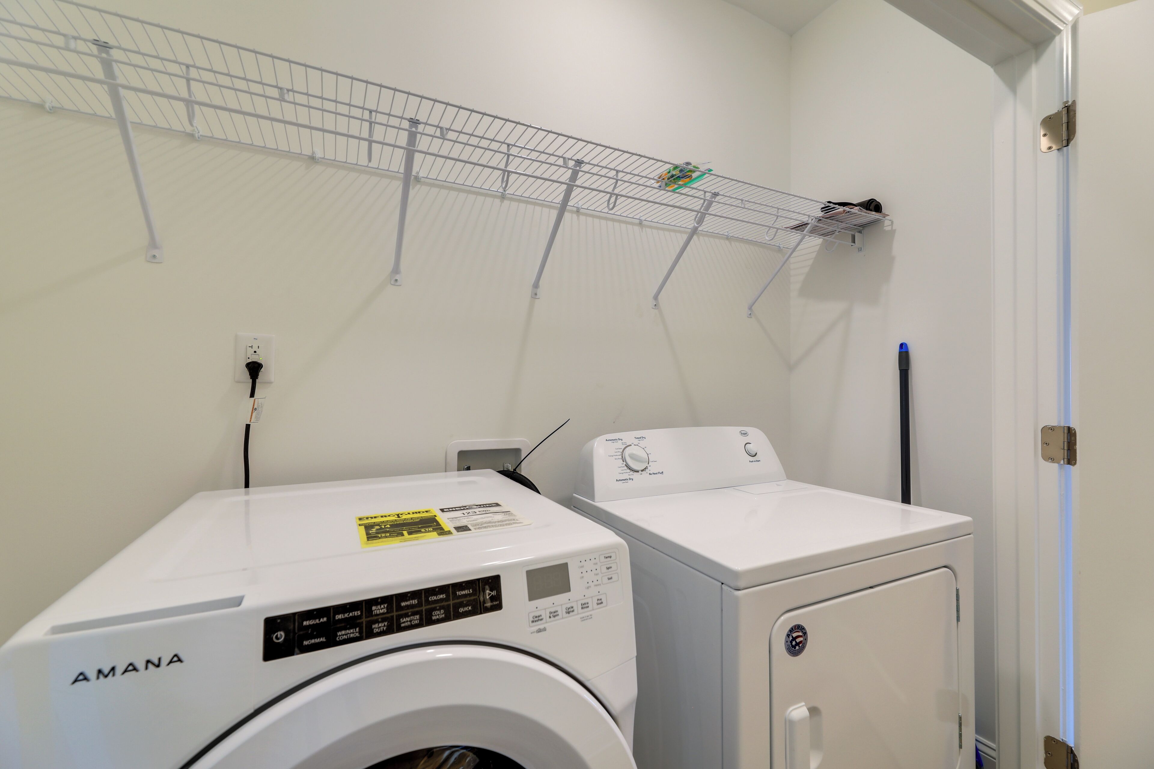 A laundry area with a front-loading washer and dryer, a metal shelf above, and a door on the right.