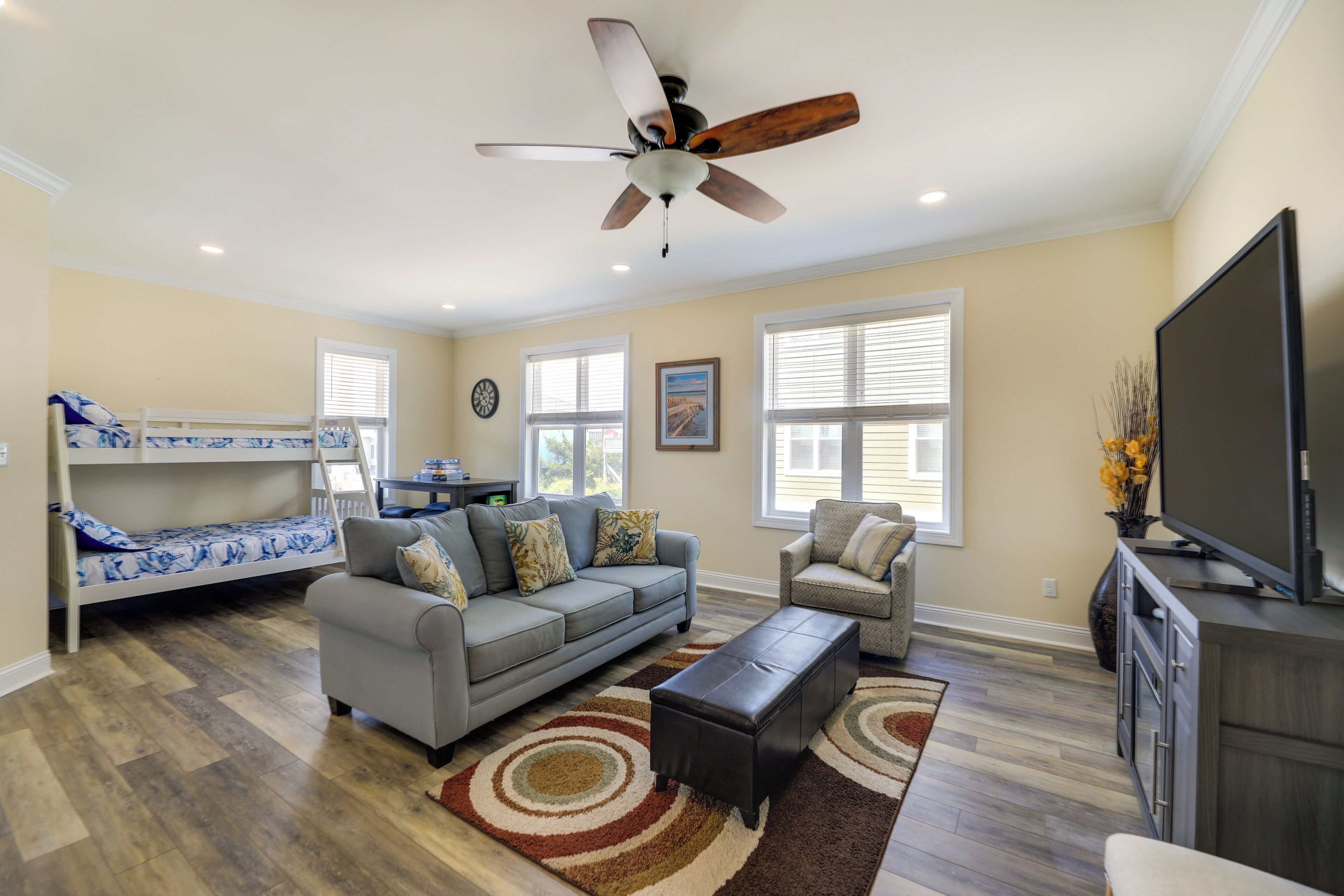 A living room with a gray sofa, a small gray armchair, a coffee table, a ceiling fan, a TV, a bunk bed, and large windows.