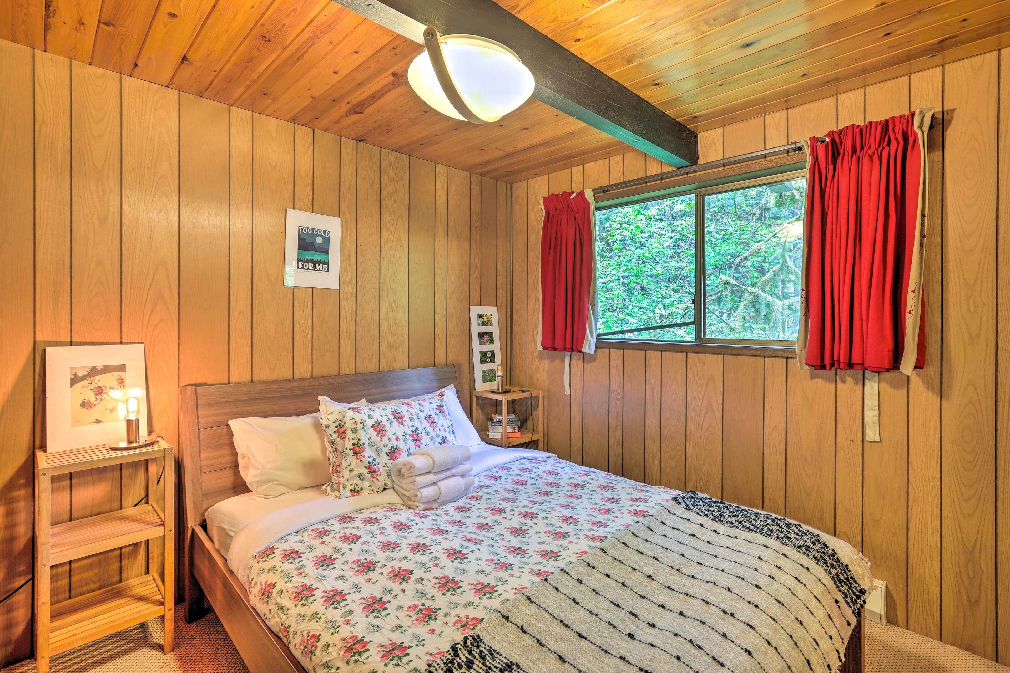 A wooden-paneled bedroom with a bed, a bedside table, a framed picture, and a window with red curtains.