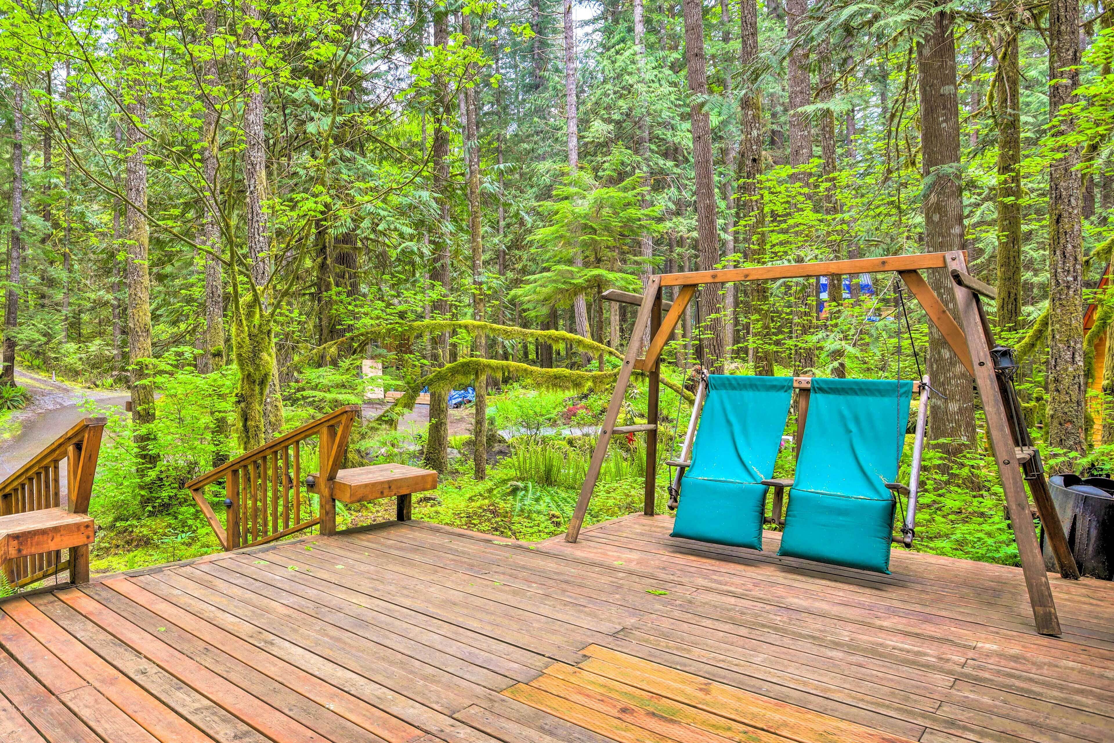 Eine Holzterrasse mit Schaukel und Bänken, umgeben von dichtem Wald.