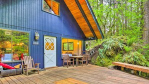 Ein blaues Holzhaus mit einer Terrasse, einem Essbereich mit Stühlen und einem Tisch sowie Blick auf den Wald.