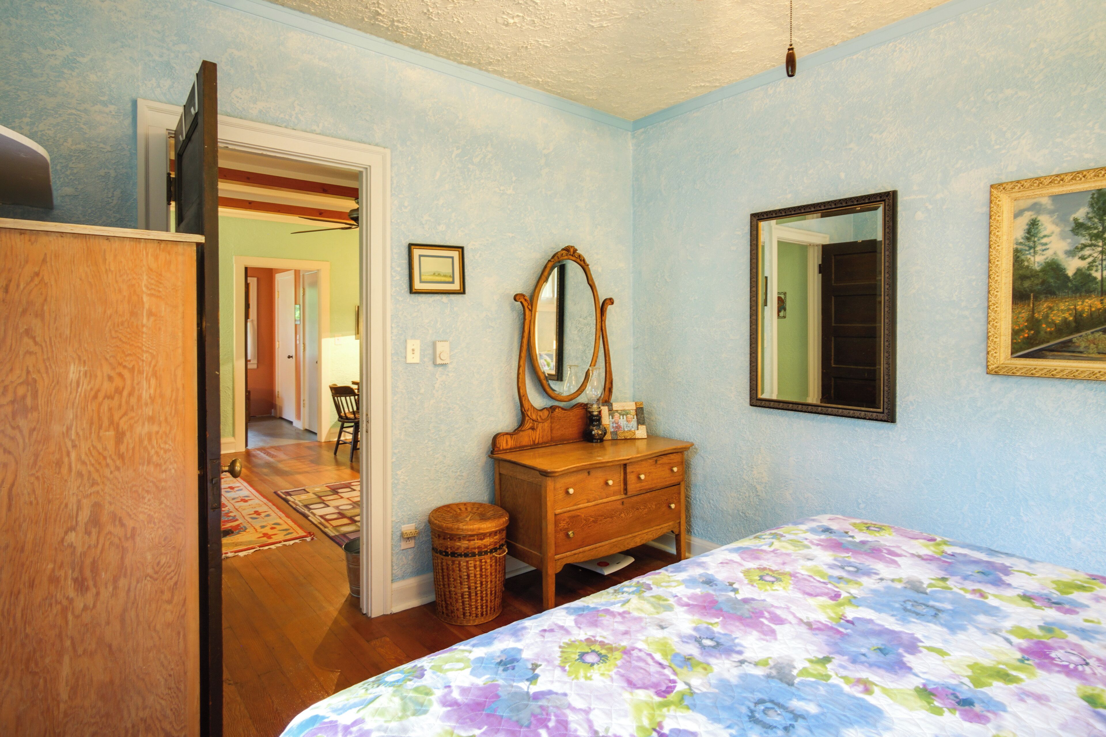 A bedroom with a wooden dresser, a bed with a floral bedspread, a mirror, and a painting on the wall.
