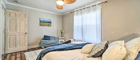 Ένα υπνοδωμάτιο με ένα κρεβάτι, έναν ανεμιστήρα οροφής, ένα παράθυρο με κουρτίνες, μια πόρτα και ένα μικρό κομοδίνο με ένα βάζο.