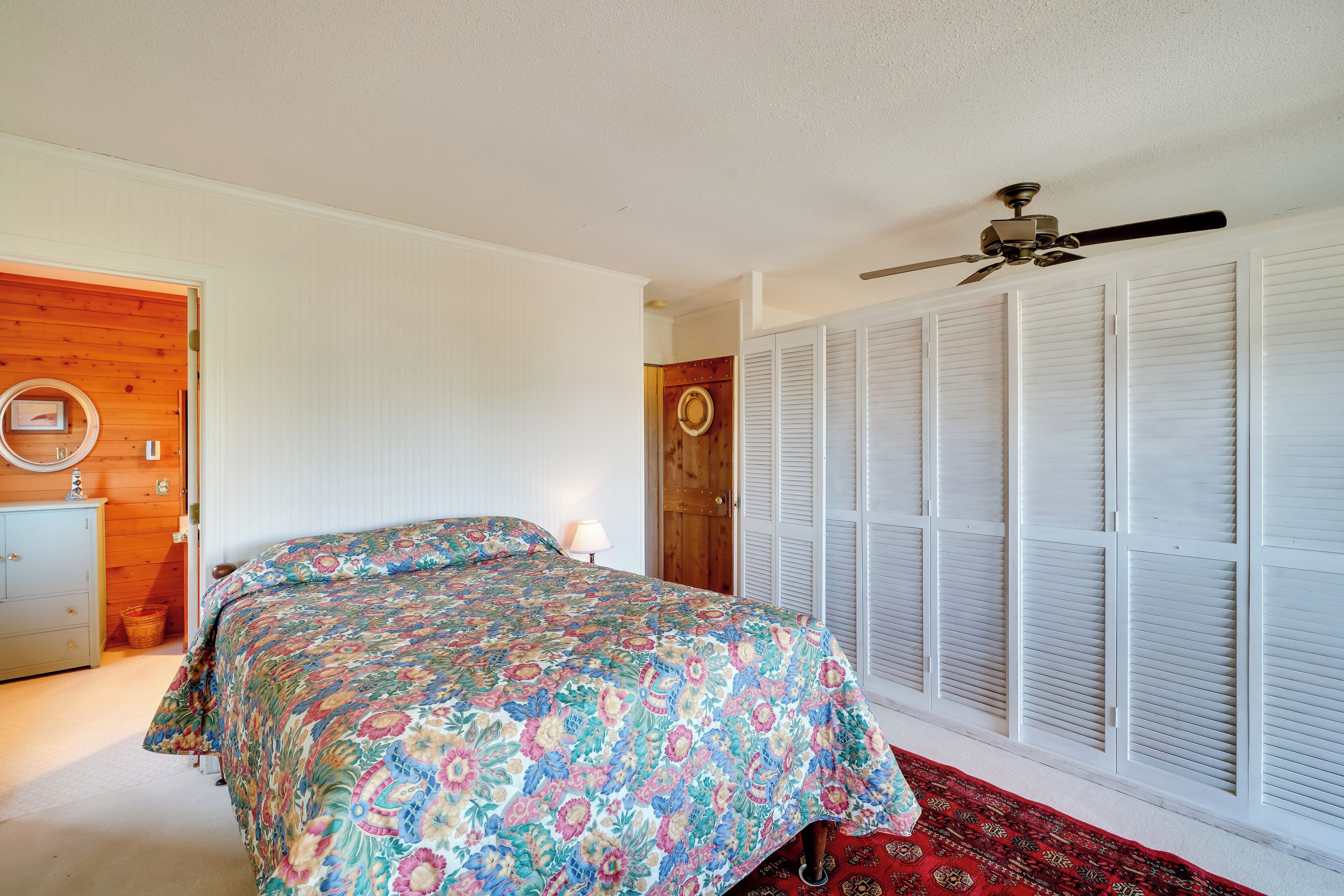 Un dormitorio con una cama, un ventilador de techo, una puerta de madera y un armario con contraventanas blancas.