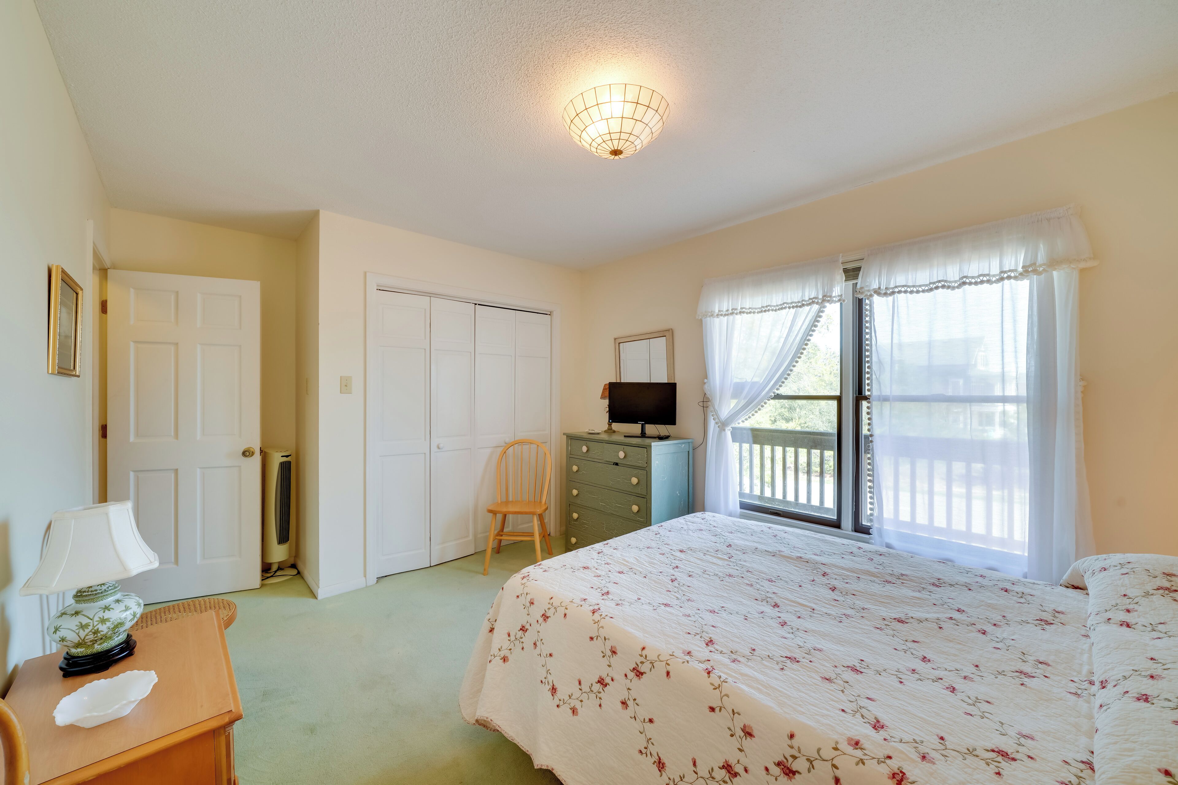 Une chambre à coucher comprenant un lit, une commode, une chaise, un bureau avec une lampe, un téléviseur et une fenêtre avec des rideaux.