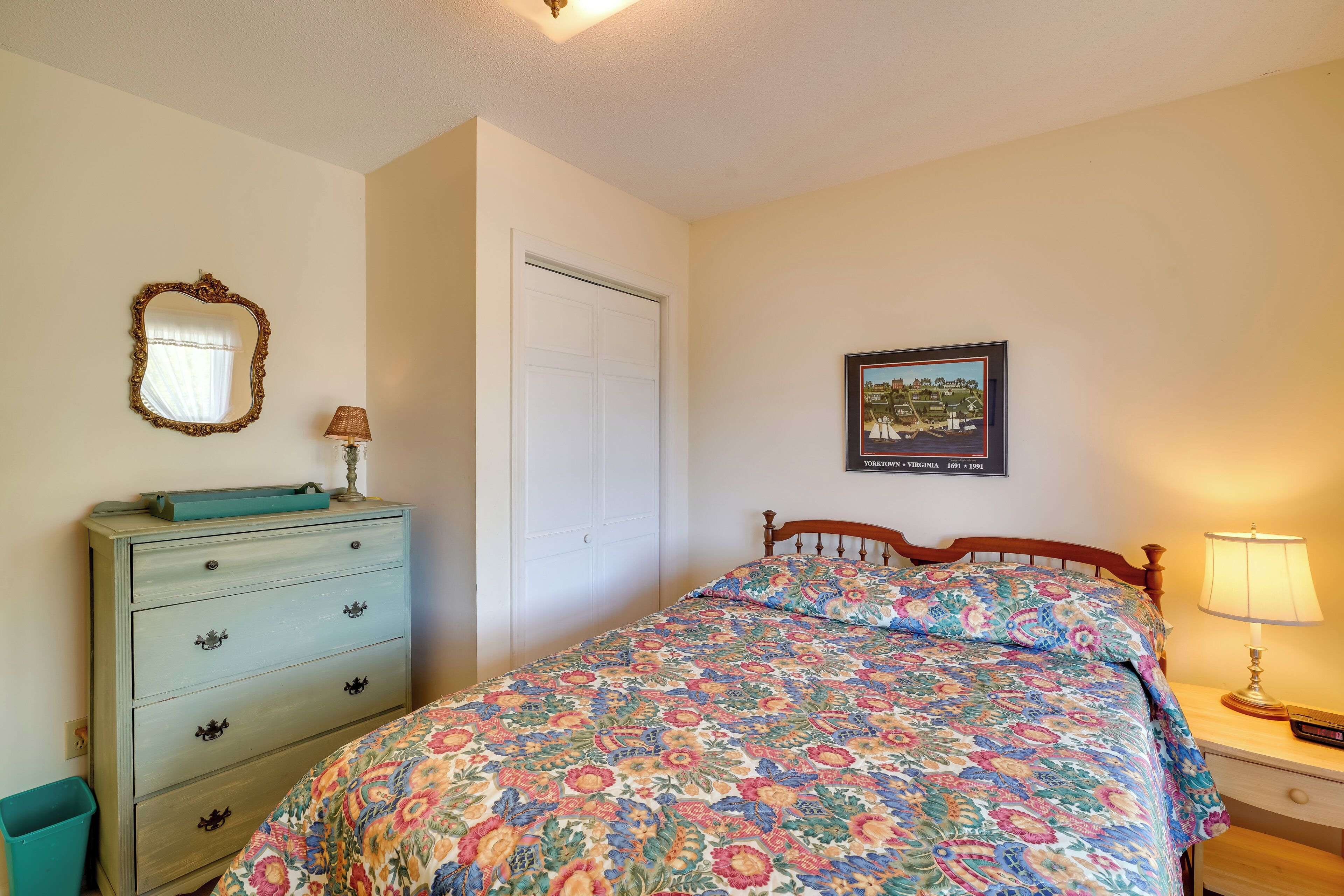 Een slaapkamer met een bed, een dressoir, een nachtkastje, een lamp en een lijst aan de muur.