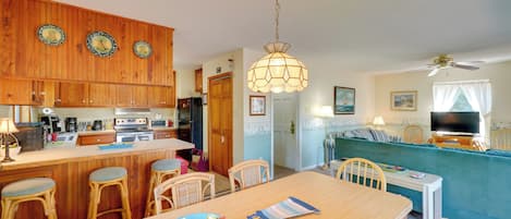 A kitchen with wooden cabinets, a central island with stools, a dining table with chairs, and a living area with a sofa and television.