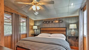 Quarto com cabeceira de madeira, ventilador de teto, cama com roupa de cama xadrez, duas mesas de cabeceira com abajures e janela com cortinas.