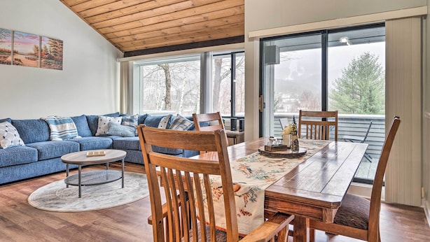 A modern living room with a wooden ceiling, a blue sofa, a dining table with chairs, and large windows offering a view of trees. - Lincoln Escape w/ Deck: 1 Mi to Loon Mountain! (Lincoln)