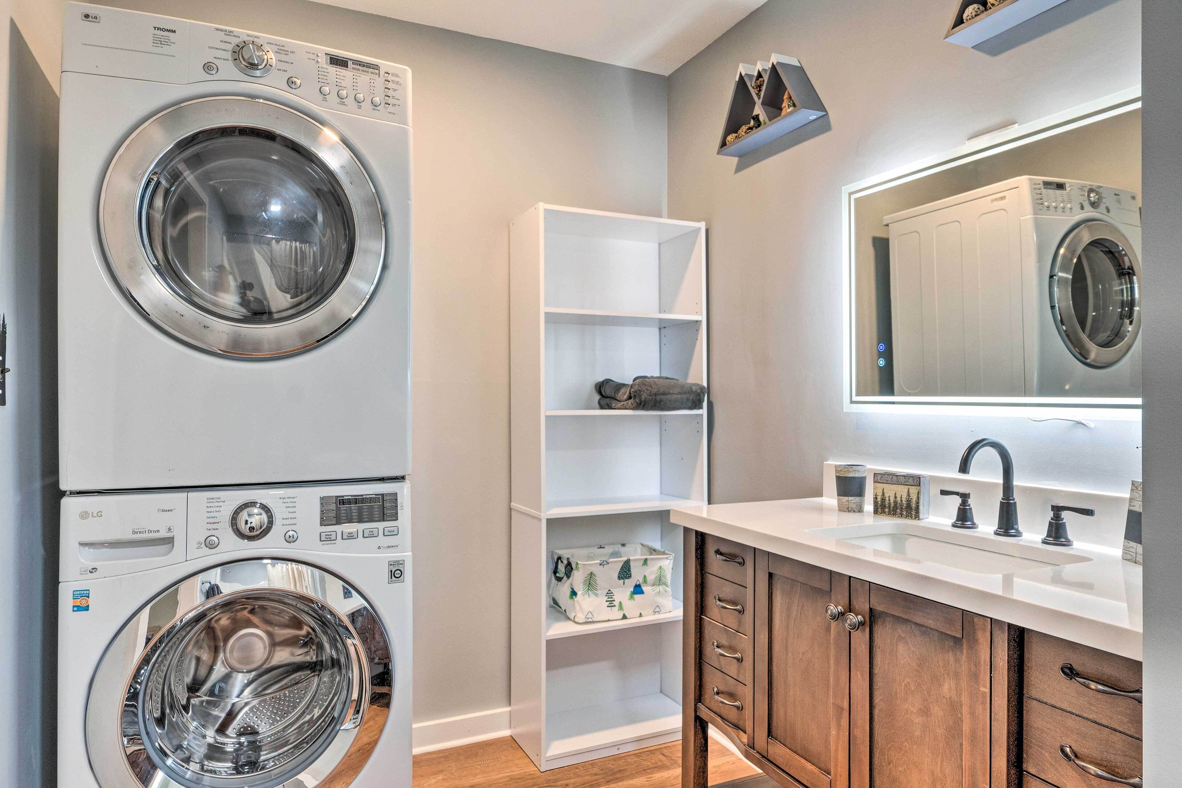 A laundry area with stacked washing machines, a white shelf unit, a wooden cabinet, and a large mirror above a sink.