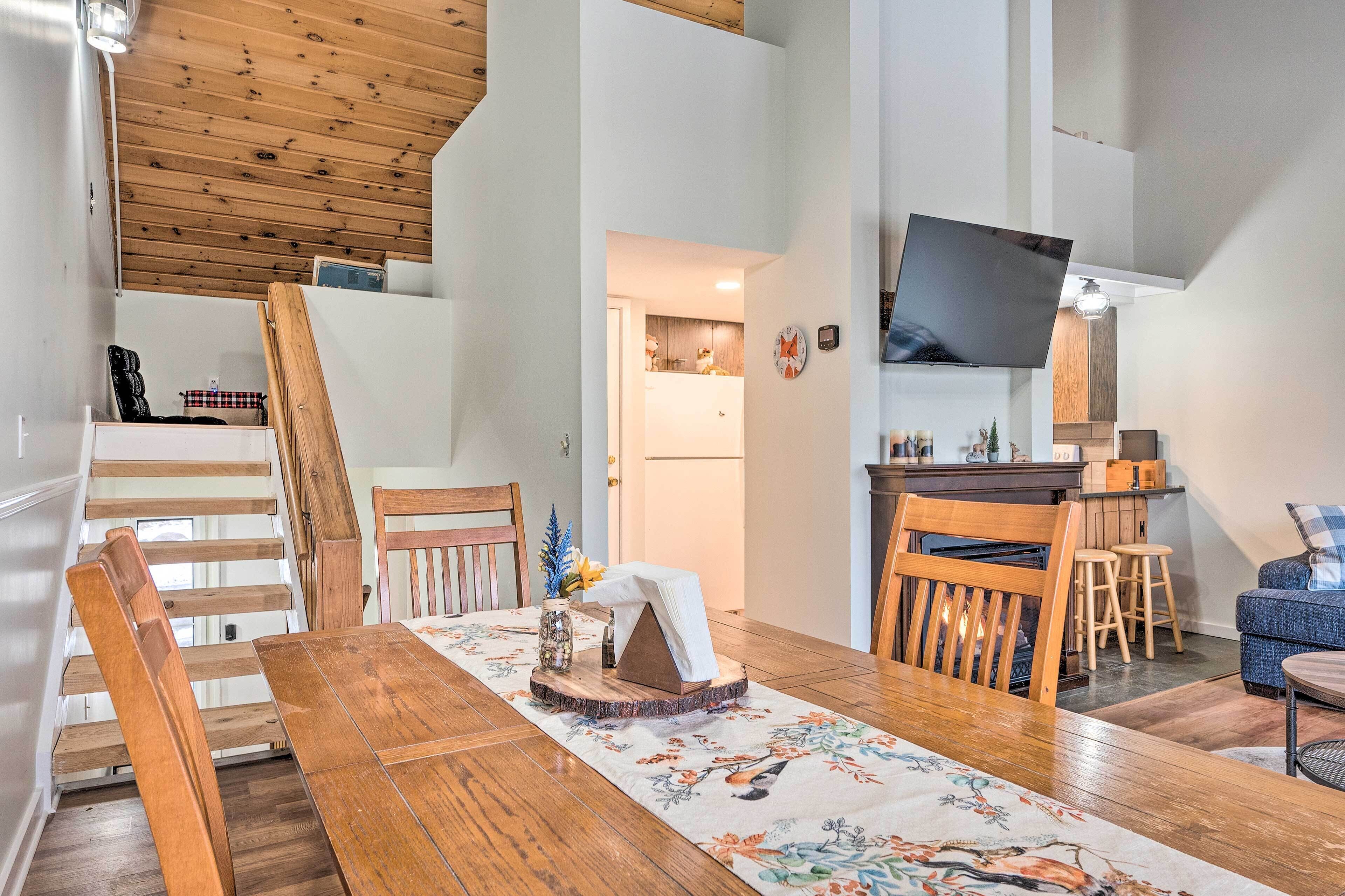 A dining area with a wooden table, chairs, and a floral table runner. A television is mounted on the wall above the table.