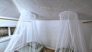 Ein Zimmer mit zwei Betten, jedes bedeckt mit einem weißen Baldachin. An der Decke sind Lichterketten angebracht.