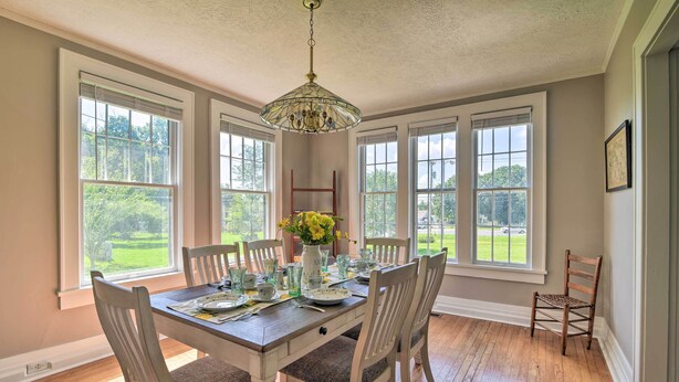 A dining area with a wooden table set for four, surrounded by white chairs, large windows with a view of greenery, and a chandelier hanging from the ceiling. - Glasgow Home w/ Porch: Close to Lake & Caves! (Glasgow)