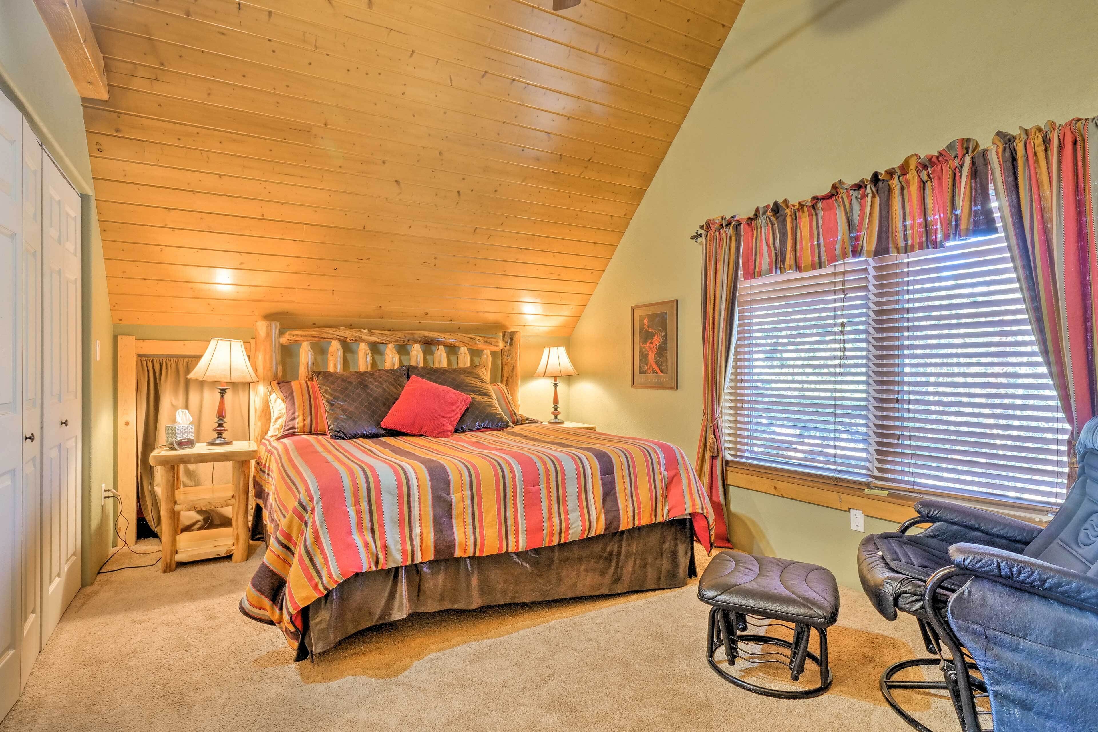 A cozy bedroom with a wooden ceiling, a large bed with striped bedding, a rocking chair, and a window with curtains.