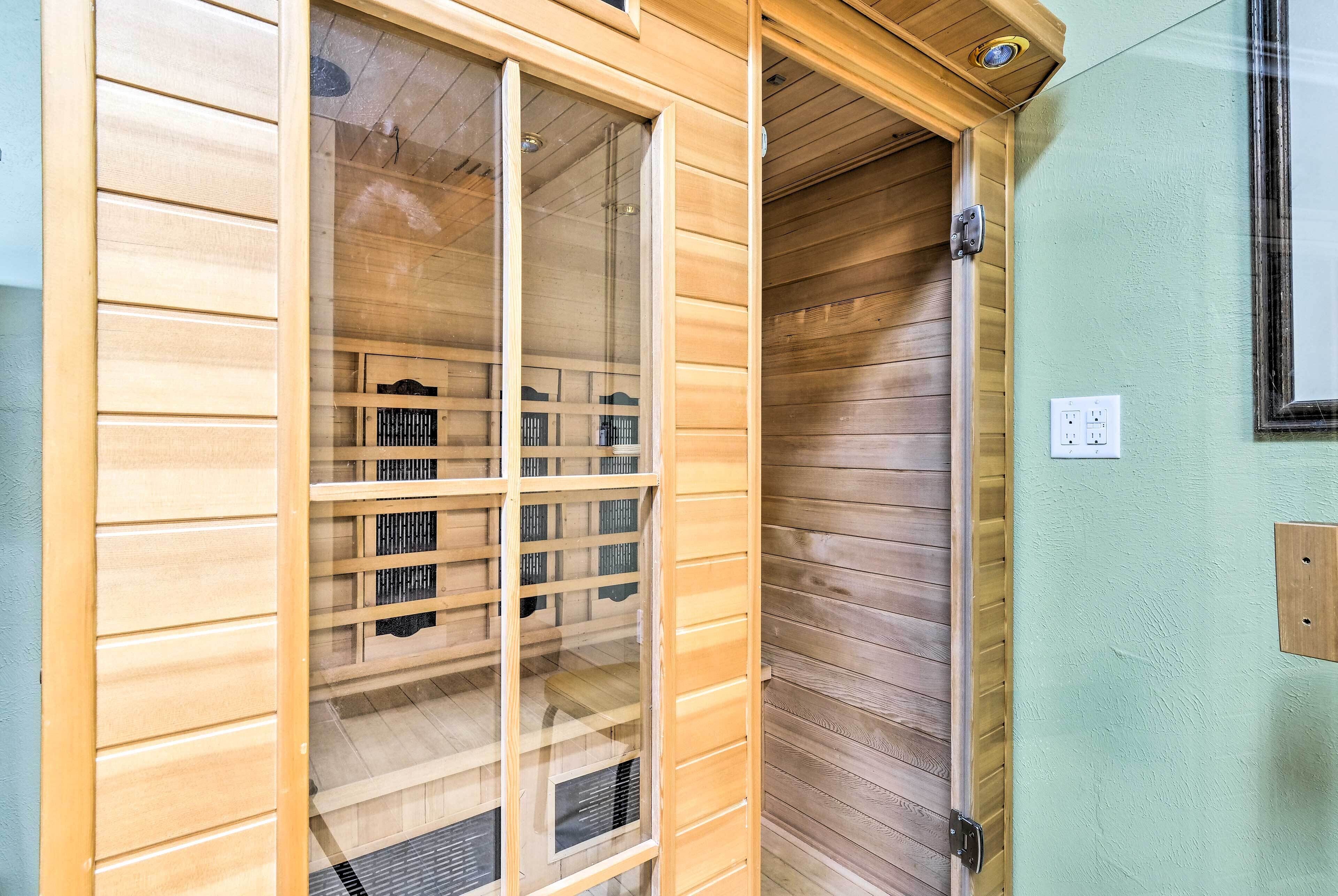 Ein Saunaraum aus Holz mit Glastür, einer hölzernen Sitzbank darin und einem Lichtschalter an der Wand.
